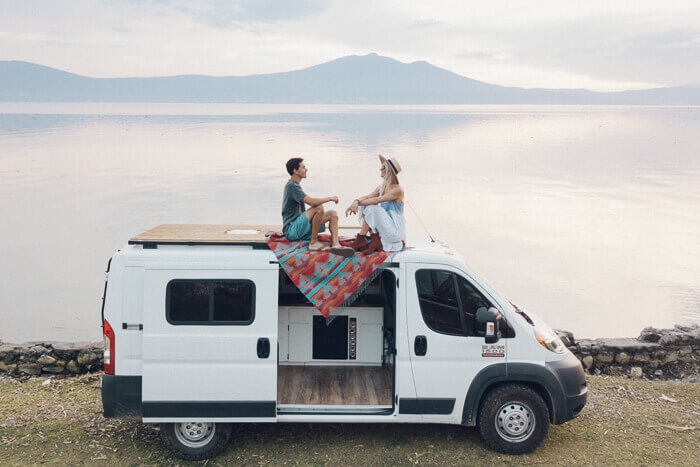 a couple sit on top of a self build campervan in front of the sea