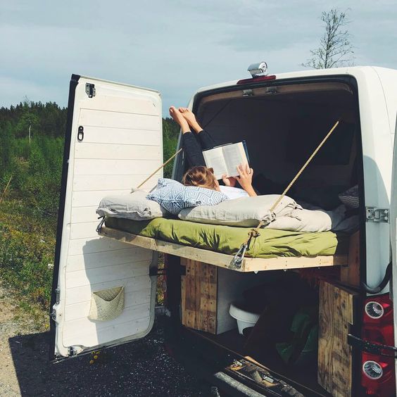 a woman reclining on a fold out campervan bed reads a book in the sunshine