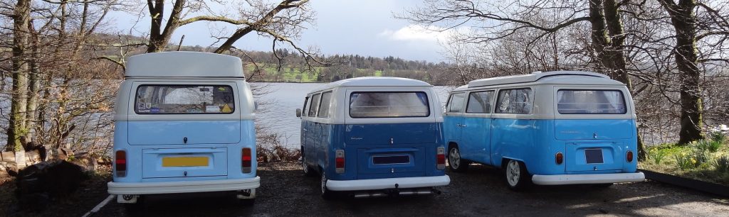 Three classic blue VW campervans in front of a lake
